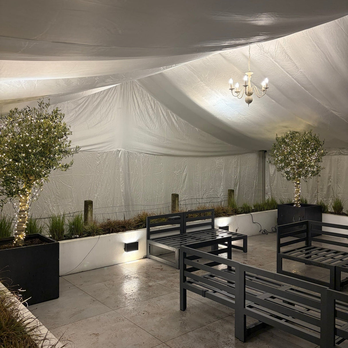 Outdoor seating area with tables and chairs on a stone patio, illuminated by string lights.