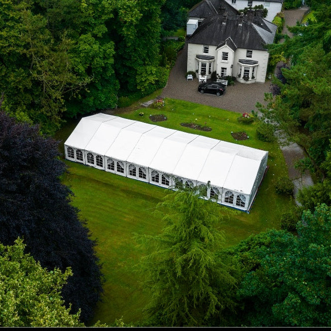 White 9x33 meter marquee tent on a grassy area surrounded by trees and a house in the background