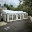 White marquee tent set up outdoors on a driveway with trees and a house in the background.